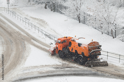 Orange snow plow truck or street sweeper machine cleans the road surface from snow. February snowfall