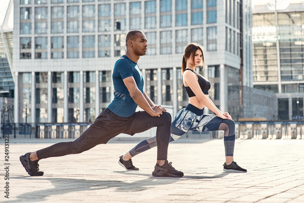 Fototapeta premium Stretching after great workout. Side view of sporty and healthy couple stretching legs during morning workout against modern buildings.