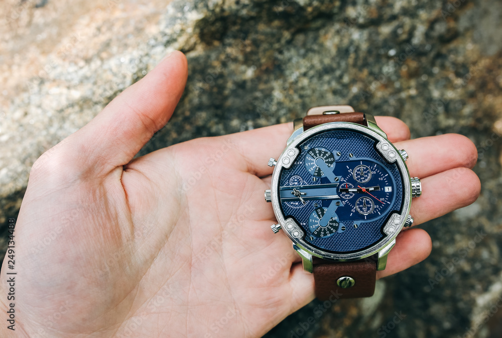 Guy is holding fashionable elegant men's watch in hands. Stylish accessories in the forest on stone. Copy space place. Guy is holding fashionable elegant men's watch in hands. Stylish accessories in the forest on stone. Copy space place.