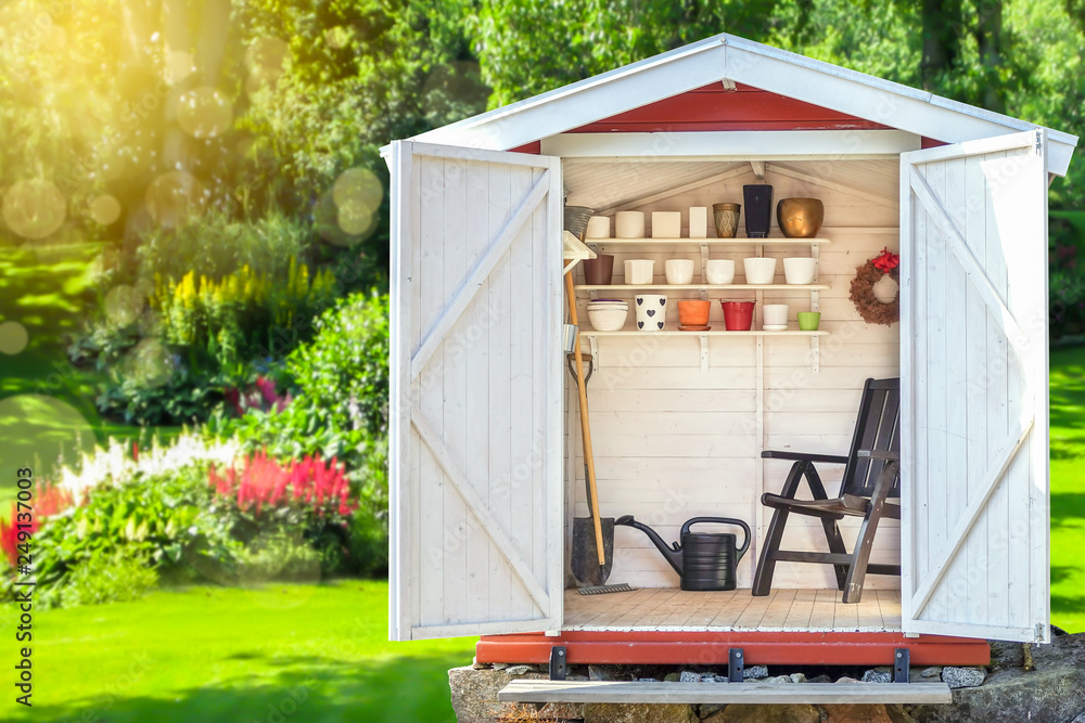 Garden shed filled with gardening tools. Shovels, rake, pots, water pitcher in storage hut. Green sunny garden in the background. - obrazy, fototapety, plakaty 