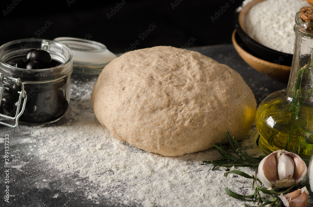 Fototapeta premium Ingredients for Focaccia: dough, rosemary, sea salt, olives, garlic, olive oil on a black wooden table.