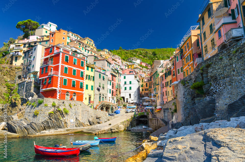 Riomaggiore traditional typical Italian fishing village in National park Cinque Terre with colorful multicolored buildings houses on hill and boats on water, clear blue sky background, Liguria, Italy