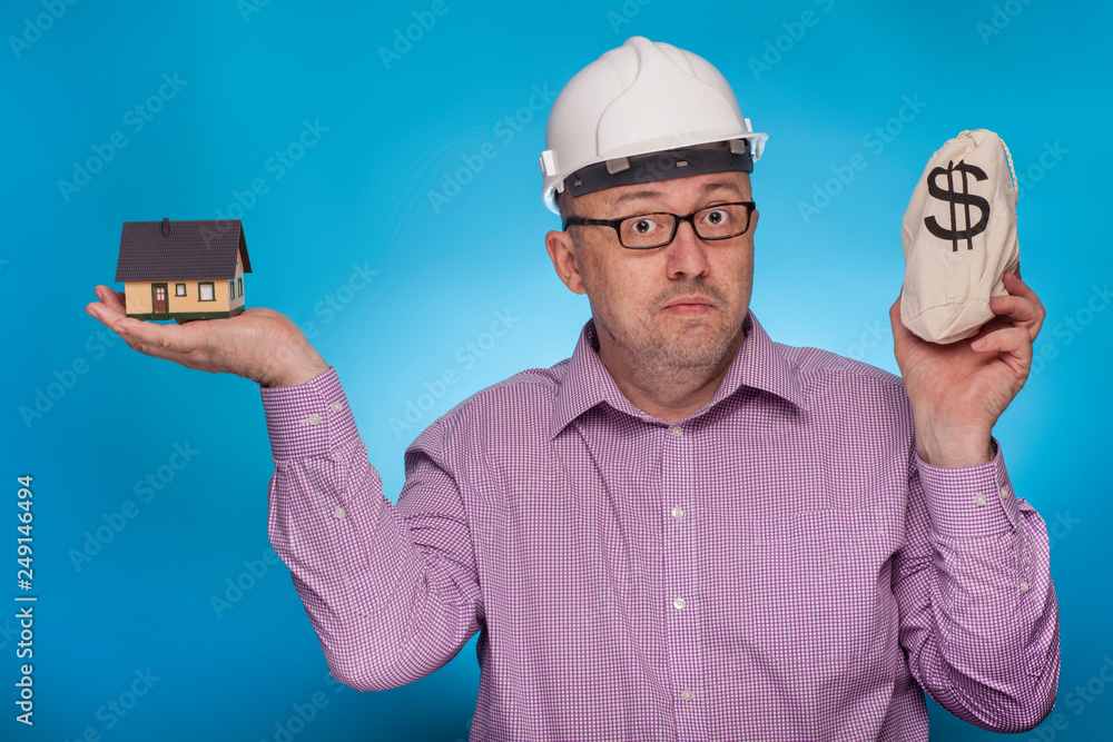 A businessman, architect in a piked shirt and hard hat has a house and a bag of dollars in his hand A businessman, architect in a piked shirt and hard hat has a house and a bag of dollars in his hand