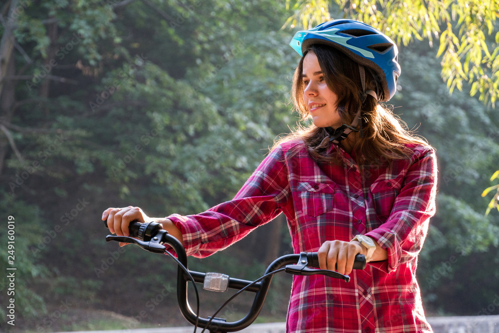 Naklejka premium Subject ecological mode of transport bicycle. Beautiful young kasazy woman wearing a blue helmet and long hair poses standing next to an orange-colored rental bike with a basket in a city park