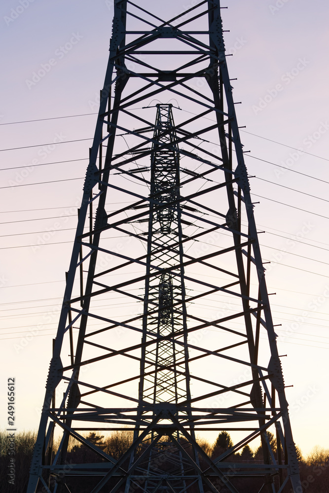 Three electricity pylons are staggered directly behind each other as silhouettes in front of a colored evening sky