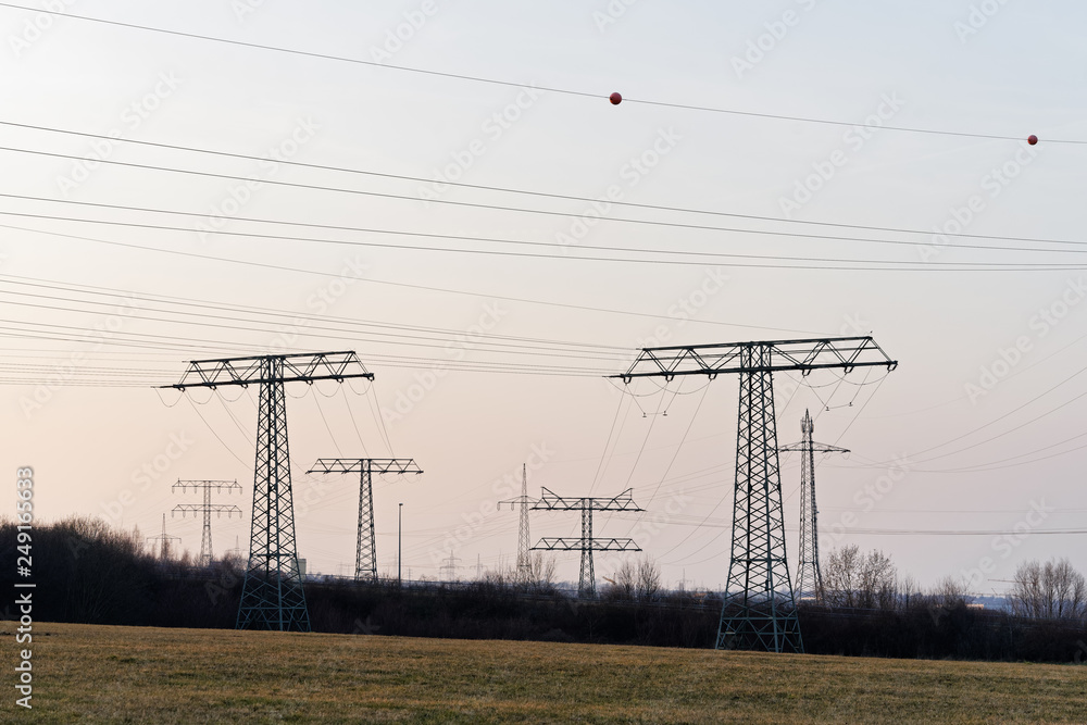 Large collection of electricity pylons in different types with cables, they stand staggered as silhouettes in front of a slightly colored evening sky in the area near a big city - Location: Germany