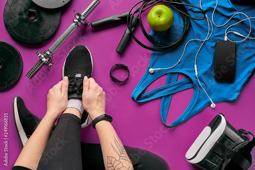 Young woman laces sneakers, preparing for training. Bottle of water, yoga mat, phone, headphones on purple background flat lay top view.