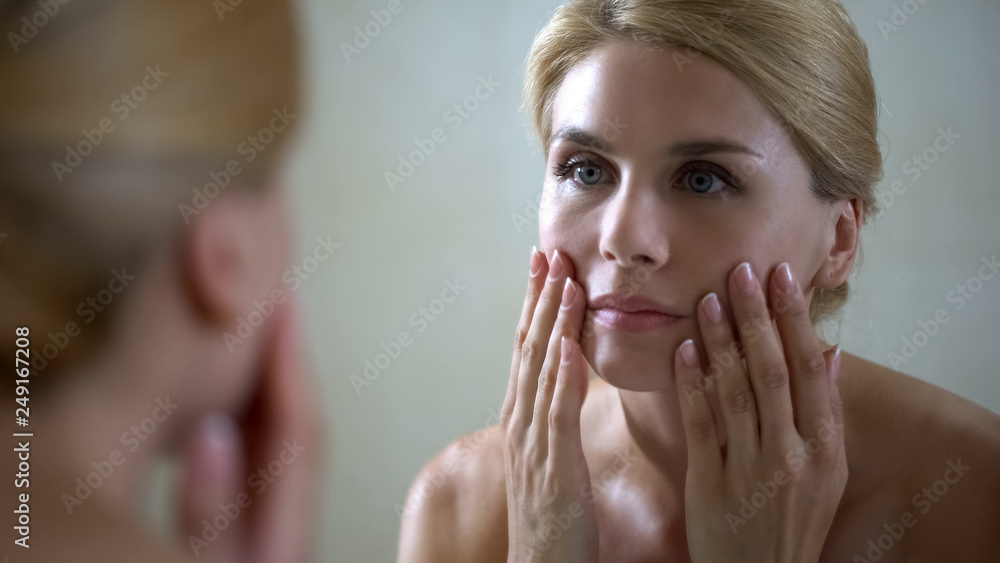 Disappointed woman looking at face reflection in mirror, anti-aging injections Disappointed woman looking at face reflection in mirror, anti-aging injections