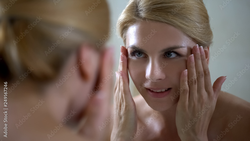 Naklejka premium Smiling woman looking in mirror, perfect beautiful skin, rejuvenating injections