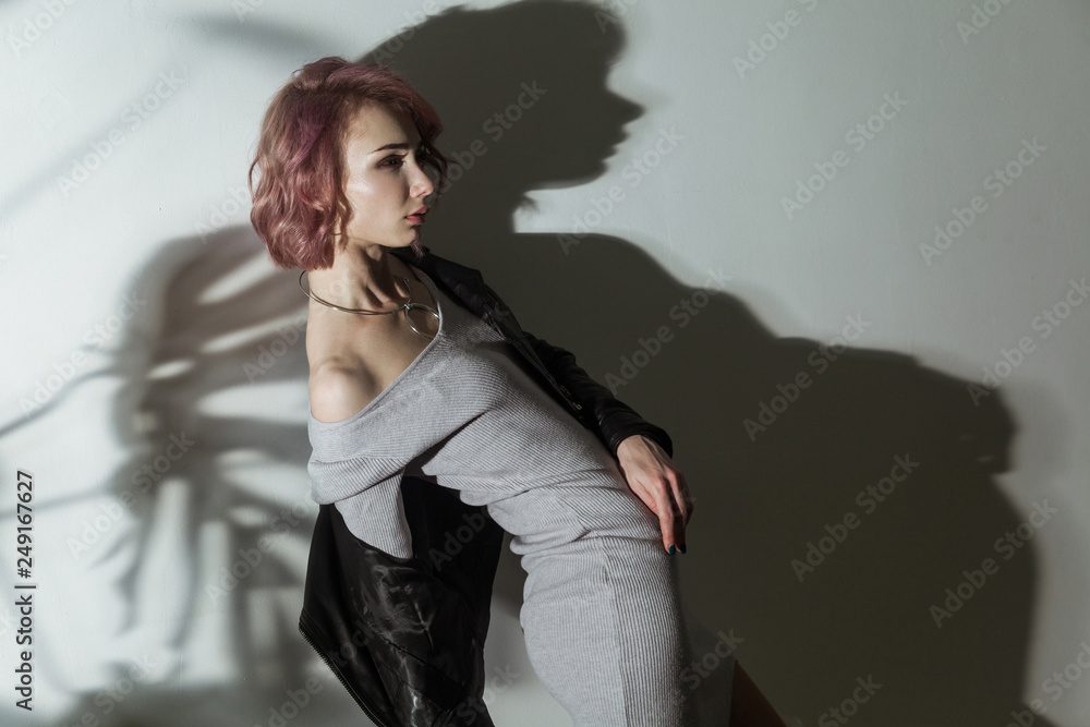 Fototapeta premium beautiful woman with makeup and short pink hair in grey dress and black leather jacket posing on grey background with dark shadow of leaf texture pattern. indoor studio shot.