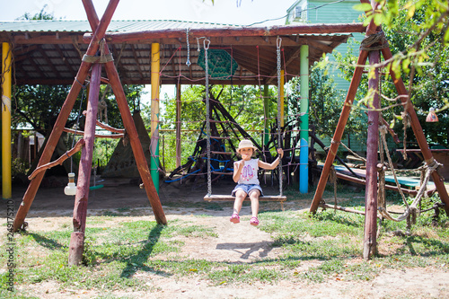 a little girl in denim shorts and a straw hat rides on a swing on a wooden Playground