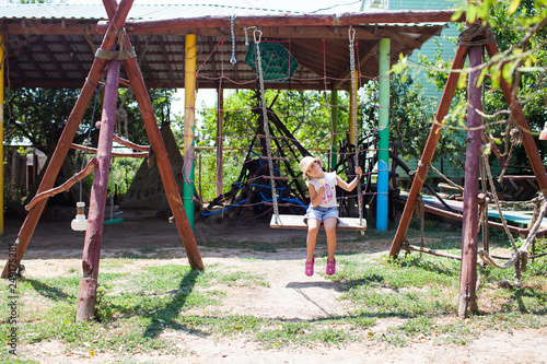 a little girl in denim shorts and a straw hat rides a swing on a wooden Playground on a Sunny day