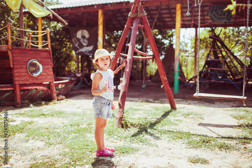 a little girl in denim shorts and a straw hat eats a Lollipop on a wooden Playground on a Sunny day