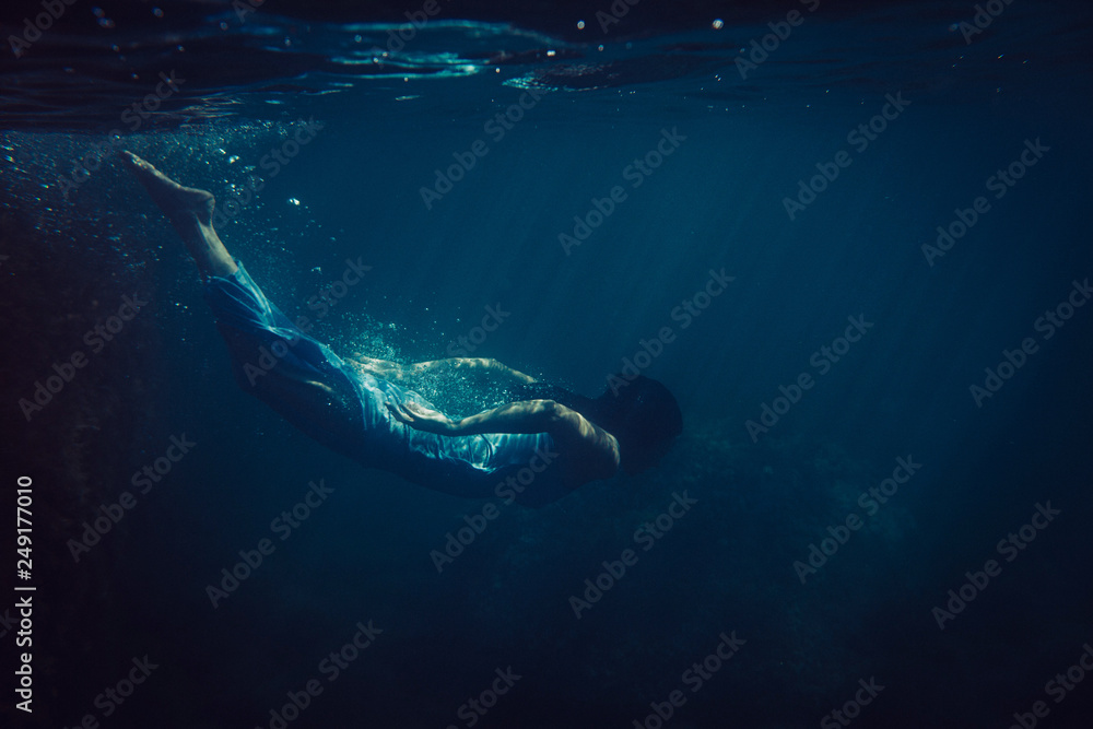 brunette girl in long blue dress dives underwater in the ocean