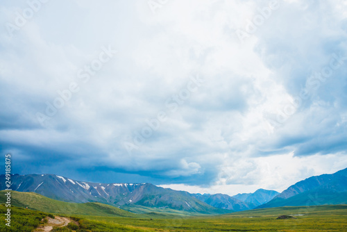 Spectacular view of distant giant mountains. Footpath through valley in highlands. Hiking path. Wonderful huge mountain range under cloudy sky. Amazing dramatic green landscape of majestic nature.