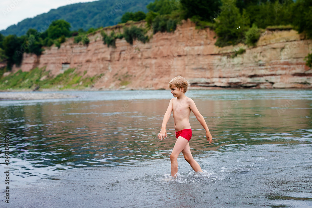 Blonde smiling boy in red swimsuit running in the mountain river Blonde smiling boy in red swimsuit running in the mountain river