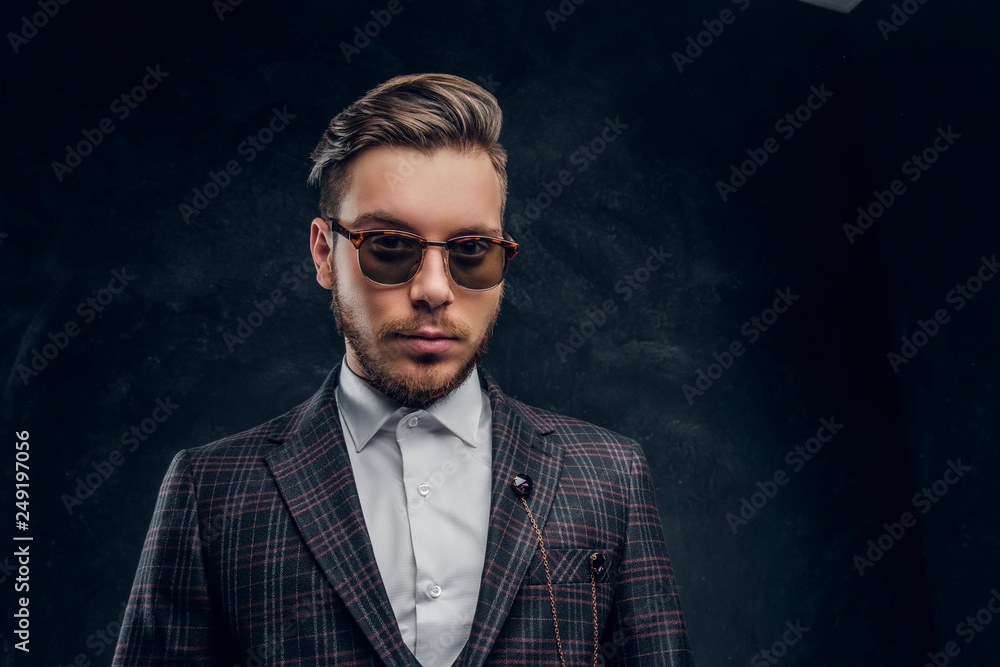 Fototapeta premium An elegantly dressed young man in sunglasses against a dark textured wall