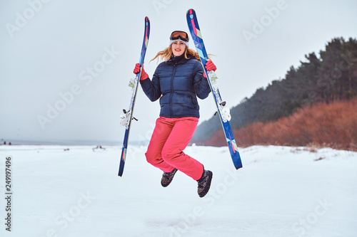 Cheerful woman wearing warming sportswear holding skis and jumping on a snowy beach