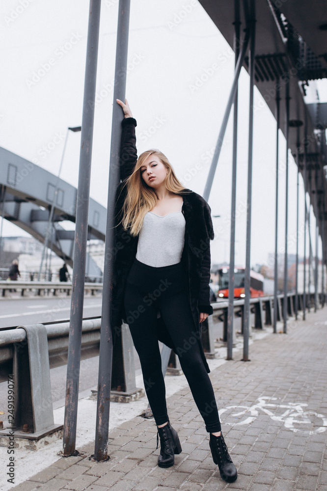 Young blondie sexy sensual girl go across the bridge in black coat. Outdoor portrait. Casual, lifestyle concept