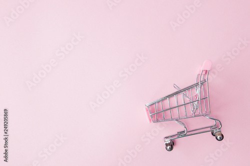 Obraz na plátne Small supermarket grocery push cart for shopping toy with wheels and pink plastic elements on pink pastel color paper flat lay background