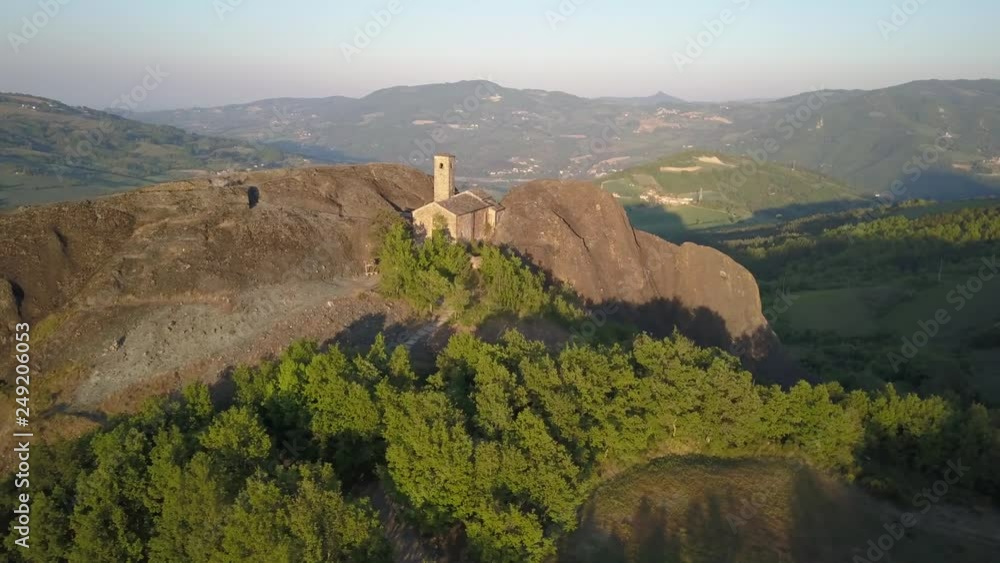 Aerial drone video at sunset time of a small ancien church built into the rocks on the hills of the Trebbia Valley, near Piacenza, Italy.
