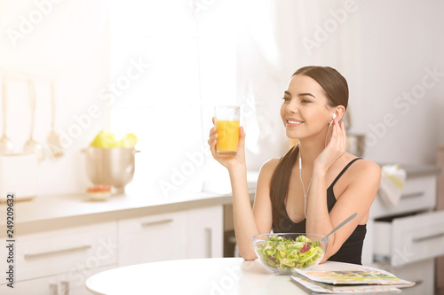 Happy young woman with glass of juice at table listening to music in kitchen, space for text. Refreshing drink