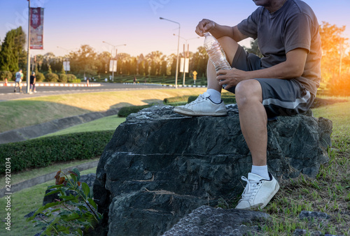 senior male sittig on stone and holding bottle of water for drinking while exercise at park outdoor with sun light background. - Image photo