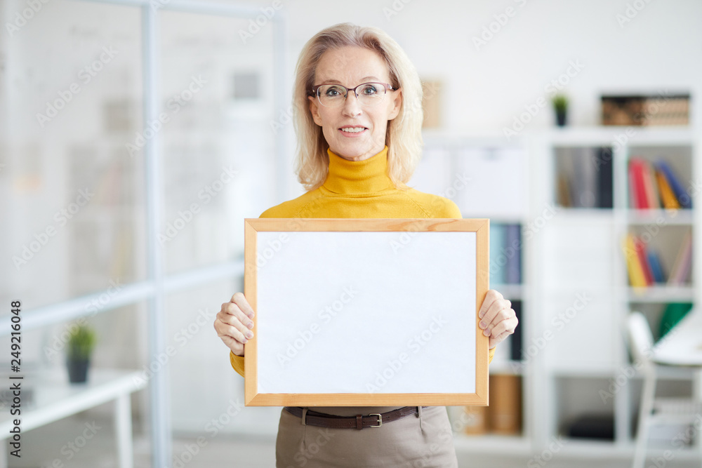 Waist up portrait of smiling mature businesswoman holding blank sign while posing in modern office, copy space Waist up portrait of smiling mature businesswoman holding blank sign while posing in modern office, copy space