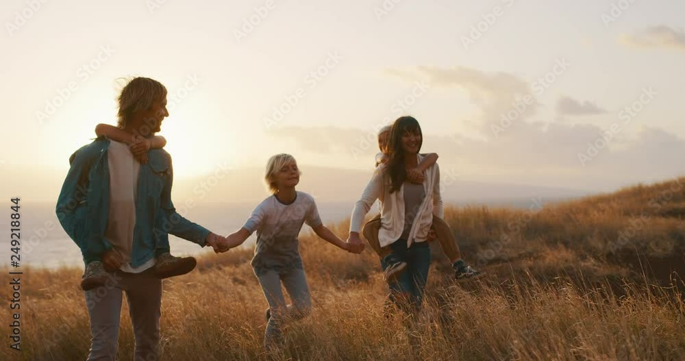 Happy smiling family holding hands walking through golden field at sunset by the ocean, piggy back ride Happy smiling family holding hands walking through golden field at sunset by the ocean, piggy back ride