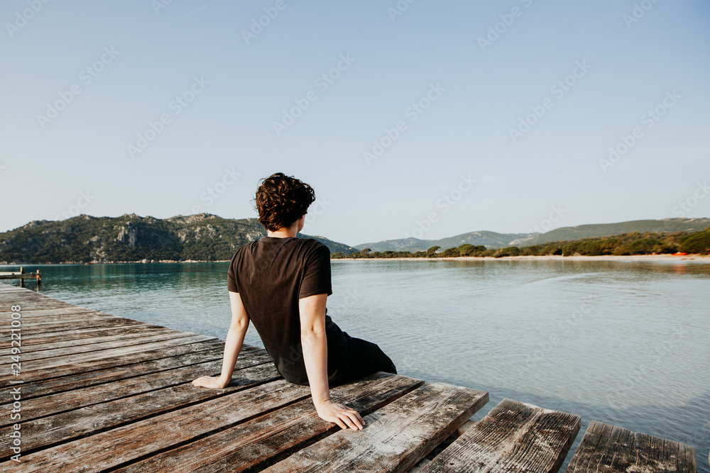 Junge Person sitzt im Sommer Urlaub auf einem Steg und genießt die Aussicht auf das Wasser Meer im Sommer