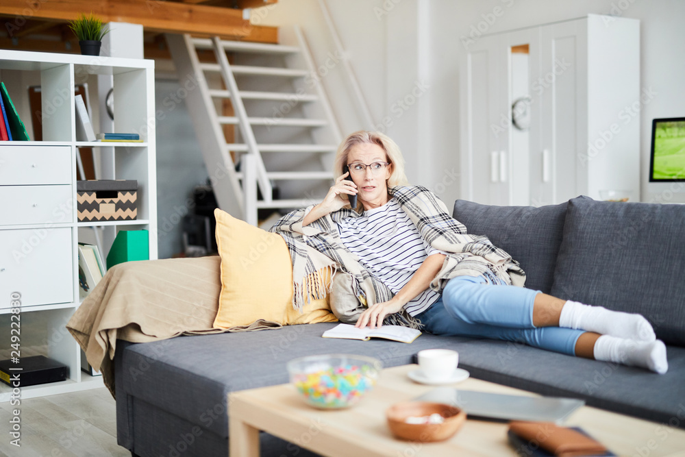 Full length portrait of contemporary woman speaking by phone while relaxing on comfortable sofa at home, copy space