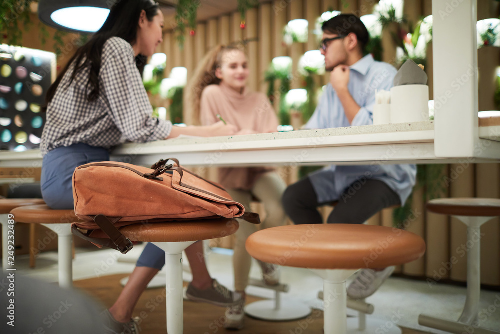 Brown backpack of college student on chair on background of friends discussing home assignment