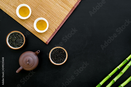 Chinese tea ceremony concept. Tea pot, tea cup, dry tea leaves, bamboo mat on black background top view copy space pattern