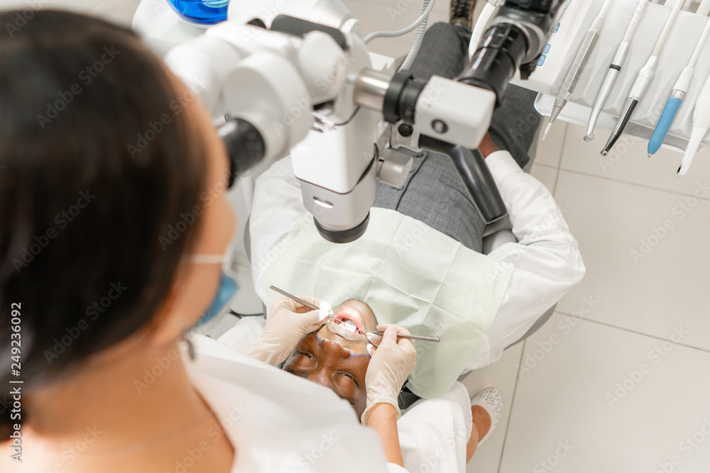 Focus on patient. Young woman dentist treating root canals using microscope in the dental clinic. Man patient lying on dentist chair with open mouth.