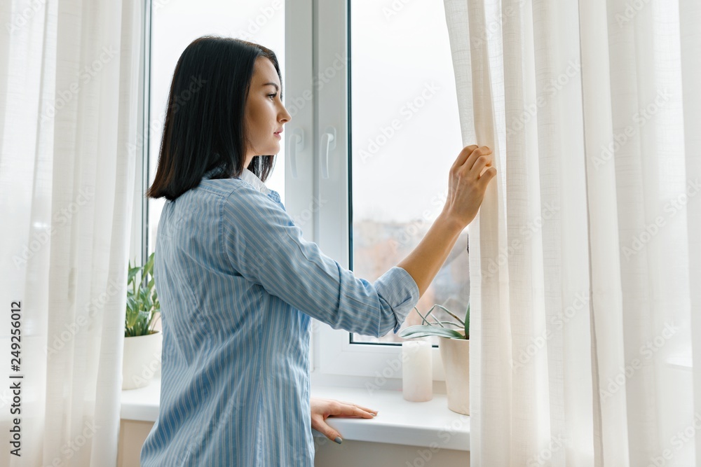 Young woman in blue shirt opening curtains looking out the window in the morning in the room, cloudy day in the city Young woman in blue shirt opening curtains looking out the window in the morning in the room, cloudy day in the city