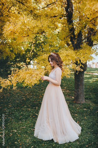 A beautiful bride in a beige dress is standing in a park with yellow leaves. The princess with a crown enjoys the fall. Wedding portrait of a cute bride with brown hair.