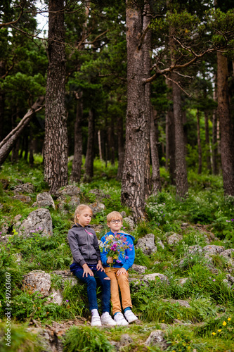Boy with glasses and blue wear with bouquet of wild flowers and blonde girl near the forest in the summer mountains. Flowers for mother and sister for the woman’s day. RUSSIA, LAGO-NAKHI, July 2018