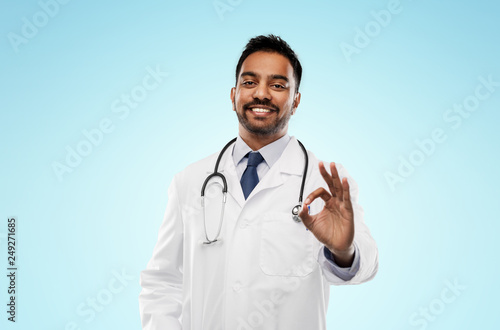 medicine, gesture and healthcare concept - smiling indian male doctor in white coat with stethoscope showing ok hand sign over blue background