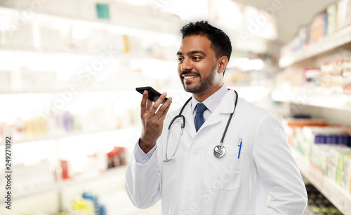 medicine, technology and healthcare concept - smiling indian male doctor or pharmacist in white coat with stethoscope using voice command recorder over drugstore background