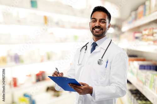 medicine, pharmacy and healthcare concept - smiling indian male doctor or pharmacist in white coat with stethoscope and clipboard over drugstore background photo