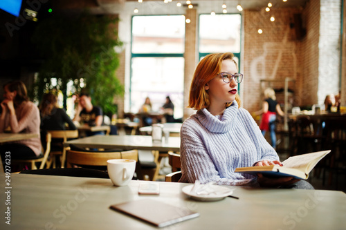 Cheerful young beautiful redhaired woman in glasses sitting at her working place on cafe and drinking coffee.