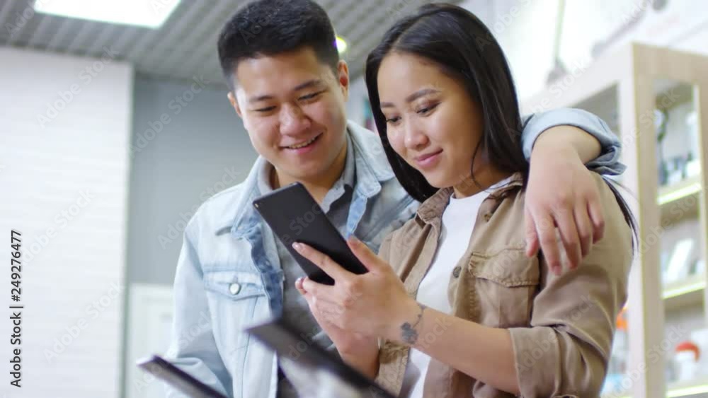 Handheld medium shot of young Asian woman testing new smartphone at gadget store and talking to boyfriend who is hugging her and smiling Handheld medium shot of young Asian woman testing new smartphone at gadget store and talking to boyfriend who is hugging her and smiling