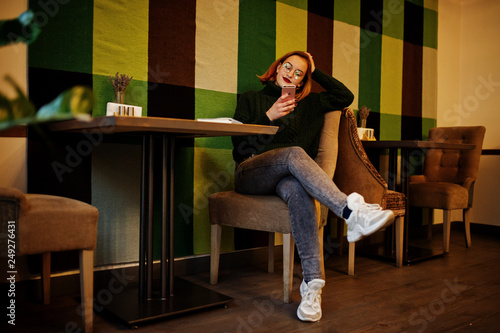 Cheerful young beautiful redhaired woman in glasses, green warm wool sweater, using her phone, while sitting at her working place on cafe.
