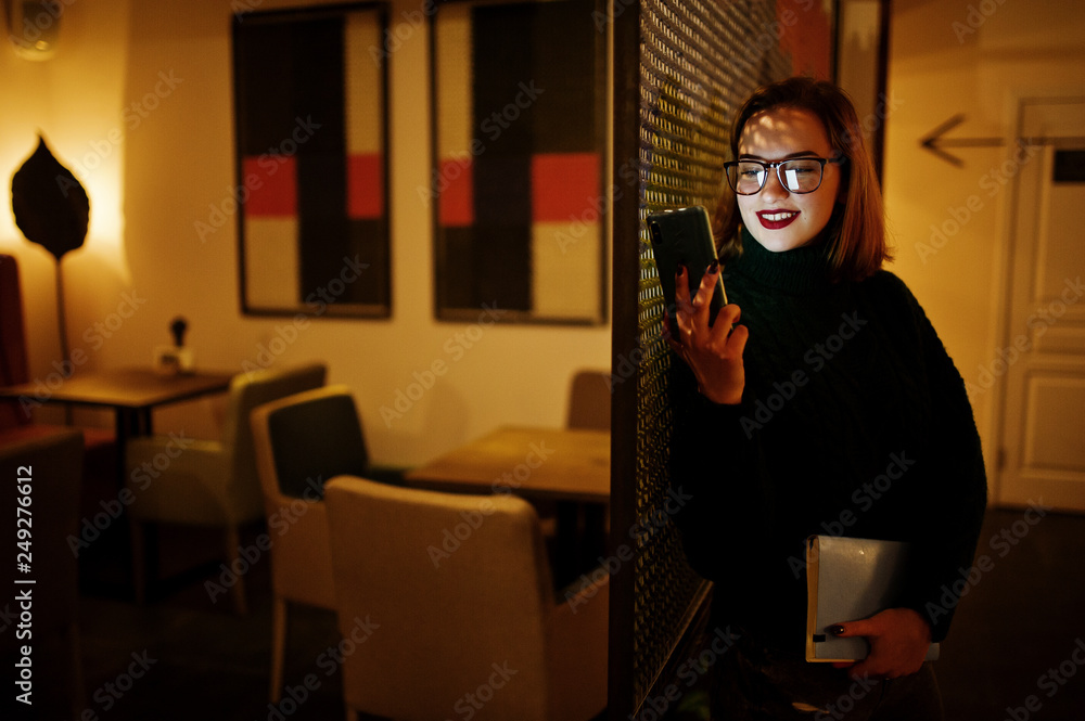 Cheerful young beautiful redhaired woman in glasses using mobile phone at dark room. Diisplaying the phone screen on her face.