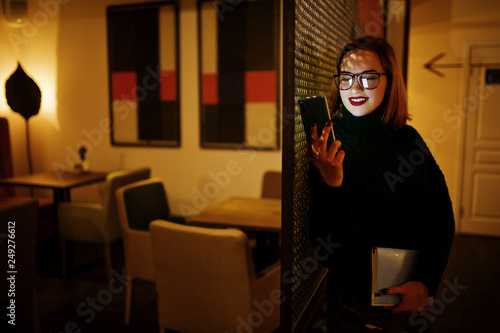 Cheerful young beautiful redhaired woman in glasses using mobile phone at dark room. Diisplaying the phone screen on her face.