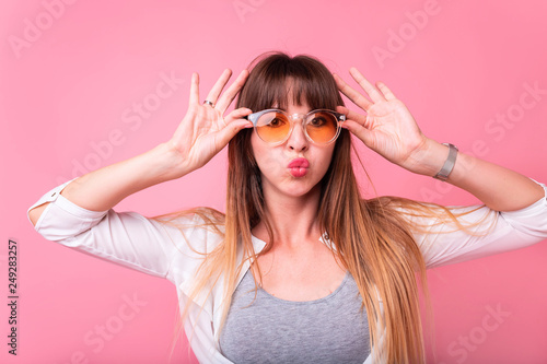 Cute lovely young woman in round sunglasses standing and sending kiss over pink background.Pleasant looking young female with straight blonde hair, keeps lips as going to kiss someone