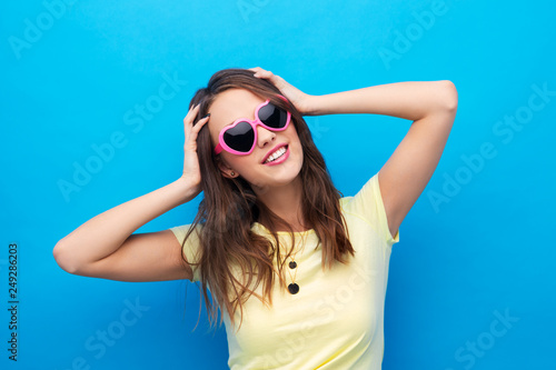 summer, valentine's day and people concept - smiling young woman or teenage girl in yellow t-shirt and heart-shaped sunglasses over bright blue background