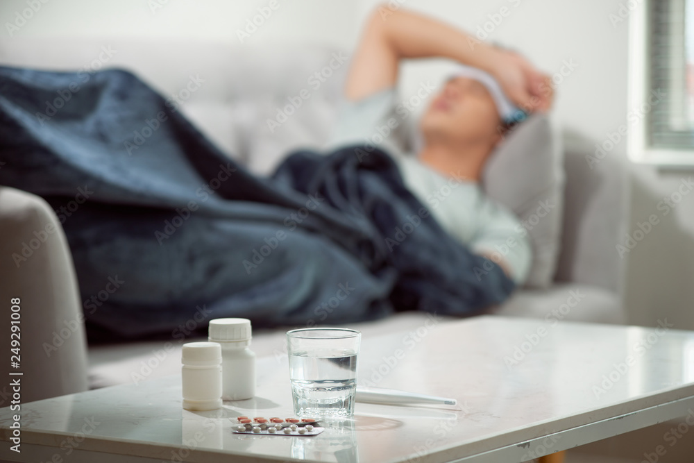 sick wasted man lying in sofa suffering cold and winter flu virus having medicine tablets in health care concept looking temperature on thermometer - obrazy, fototapety, plakaty 