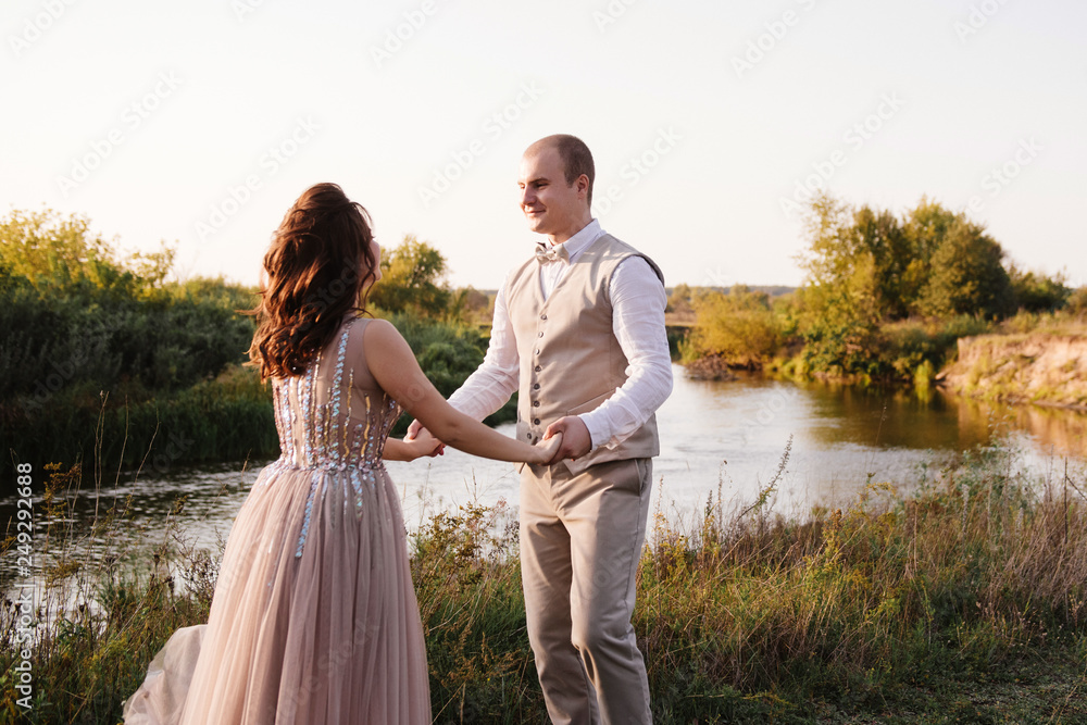 Wedding near the river in field at sunset with brown horse. Bride in light airy dress in color of dusty rose. Beige dress with sparkles. Light suit with bow tie. bride and groom embrace and kiss. Wedding near the river in field at sunset with brown horse. Bride in light airy dress in color of dusty rose. Beige dress with sparkles. Light suit with bow tie. bride and groom embrace and kiss.