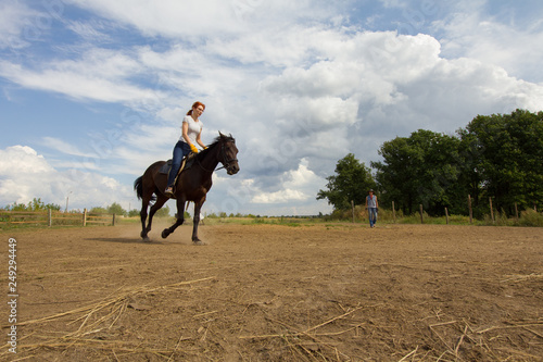 A redhead woman in white t-shirt riding a horse on the field. A green bush on a background